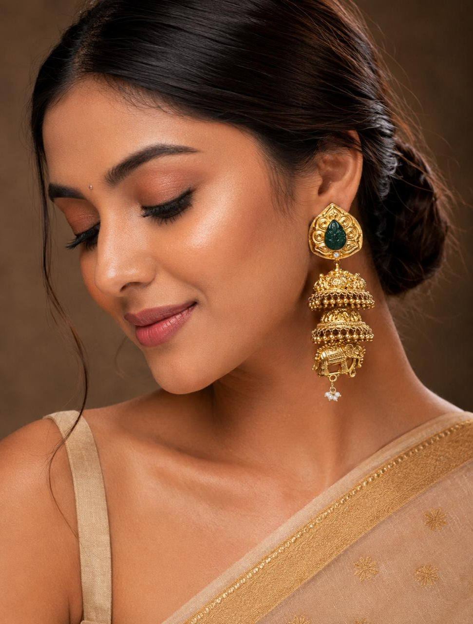 A close-up of a woman wearing an ornate gold jhumka earring with a green stone and a small white dangling accent, paired with a beige saree against a warm brown background.